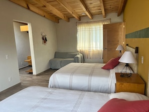 A bedroom with two beds, a wooden ceiling, a window with curtains, and a bedside table with lamps.