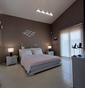 A bedroom with a bed, bedside tables, a wall-mounted light fixture, a window with blinds, and a wall decoration. - Mythos Luxury  rooftop duplex  apatment with panoramic view (Corfu)