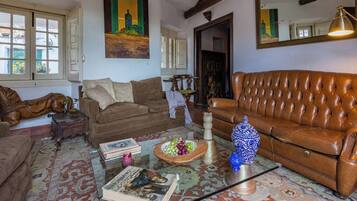 Un salon avec un canapé en cuir marron, une table basse en verre et un tapis à motifs. Il y a deux fenêtres avec des rideaux, un tableau au mur et une lampe sur une table d’appoint.