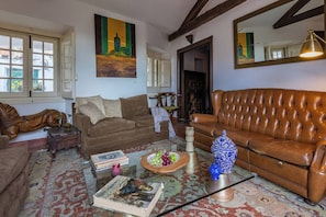 Ein Wohnzimmer mit einem braunen Ledersofa, einem Glastisch und einem gemusterten Teppich. Zwei Fenster mit Vorhängen, ein Bild an der Wand und eine Lampe auf einem Beistelltisch.