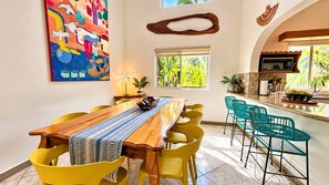 A dining area with a wooden table and yellow chairs, a kitchen with a microwave, and a colorful abstract painting on the wall. - Casa Arcos - Coastal Escape - Entire House & Attached Apartment 5 Min Beach Walk (Playa Hermosa)