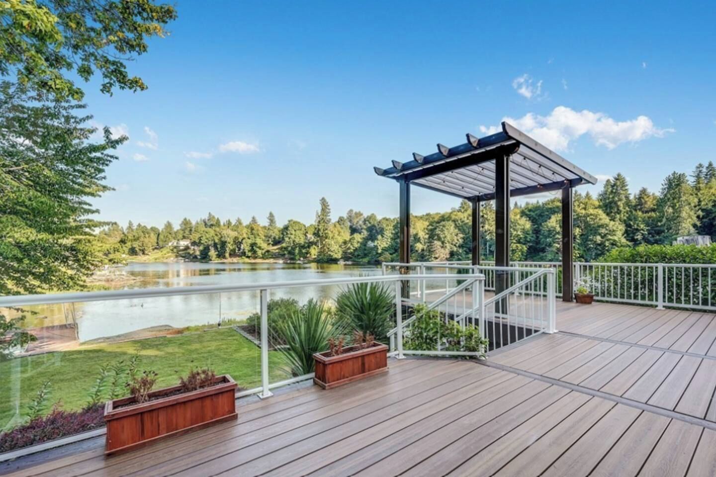 A wooden deck with a pergola, overlooking a lake and surrounded by greenery.