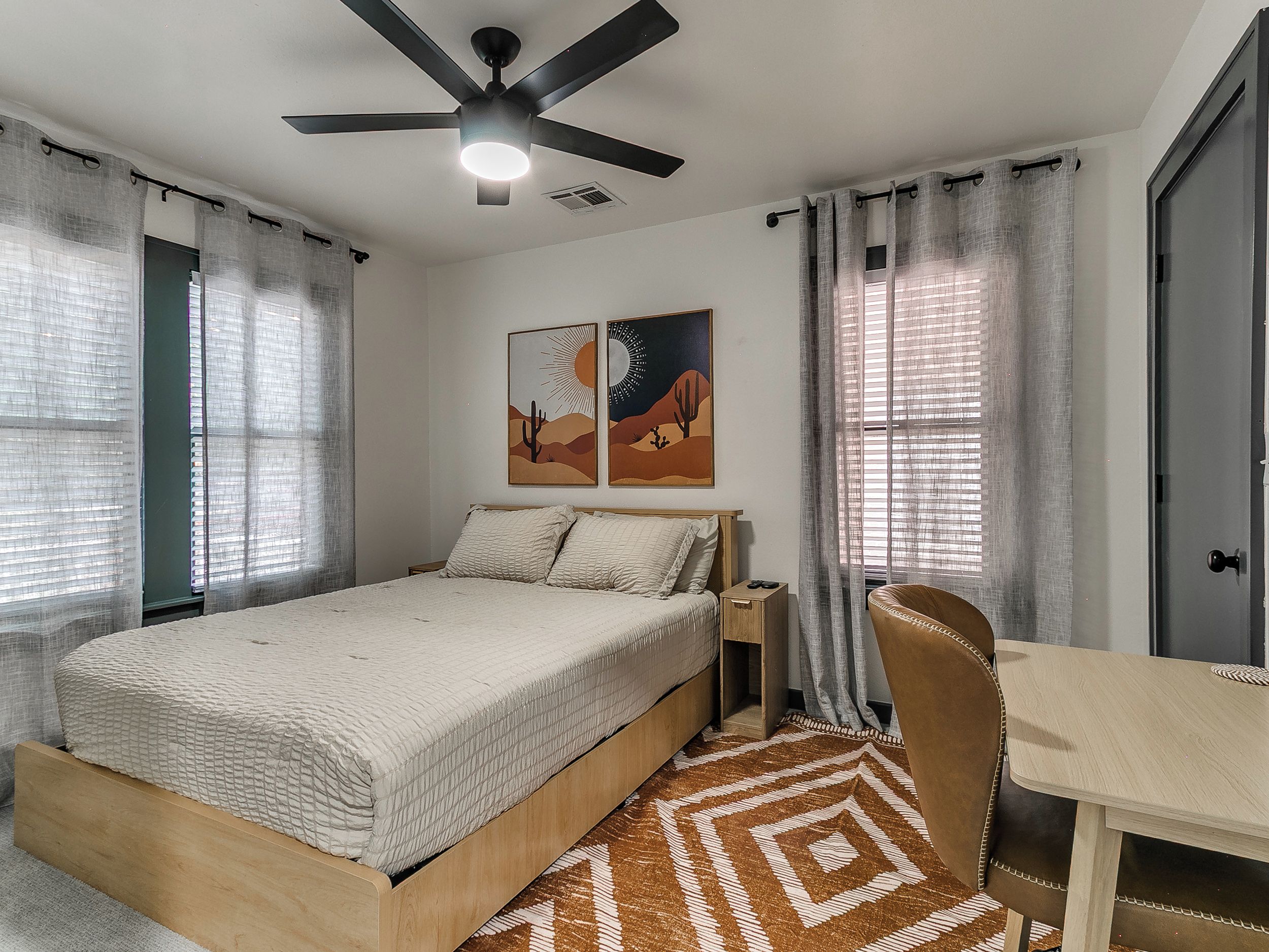 A bedroom with a bed, a desk, a chair, a ceiling fan, and artwork on the wall.