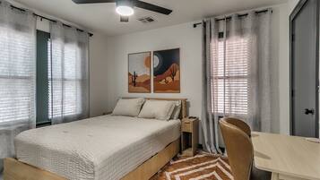 A bedroom with a bed, a desk, a chair, a ceiling fan, and artwork on the wall.