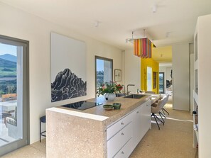 A modern kitchen with a large island, a dining area with a table and chairs, and a view of the outdoors. - Designer Villa in the Antiparos Countryside (Kampos)