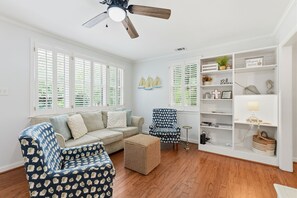 Sala de estar com sofá, poltrona estampada, mesinha lateral, estante e ventilador de teto.