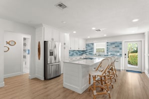 A modern kitchen with white cabinetry, a stainless steel refrigerator, a large island with bar stools, and a blue-tiled backsplash. - 2-bedroom townhome Island living Canal front Walk to the BEACH (Jensen Beach)