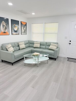 A modern living room with a light green sectional sofa, a glass coffee table, and three abstract paintings on the wall. - Spacious Black Luxe Retreat with 2 Kitchens, 2 Master Suites & Luxury Comfort (Tampa)