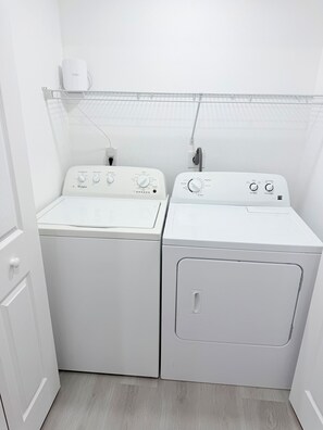 A laundry room with two front-loading washers and dryers, a power outlet, and a wall-mounted device. - Spacious Black Luxe Retreat with 2 Kitchens, 2 Master Suites & Luxury Comfort (Tampa)