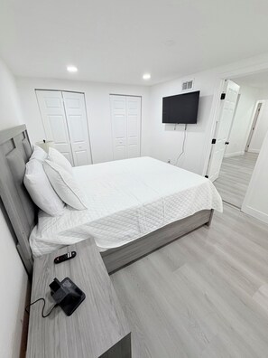 A hotel room with a bed, a TV mounted on the wall, a bedside table with a remote control, and a door leading to another room. - Spacious Black Luxe Retreat with 2 Kitchens, 2 Master Suites & Luxury Comfort (Tampa)