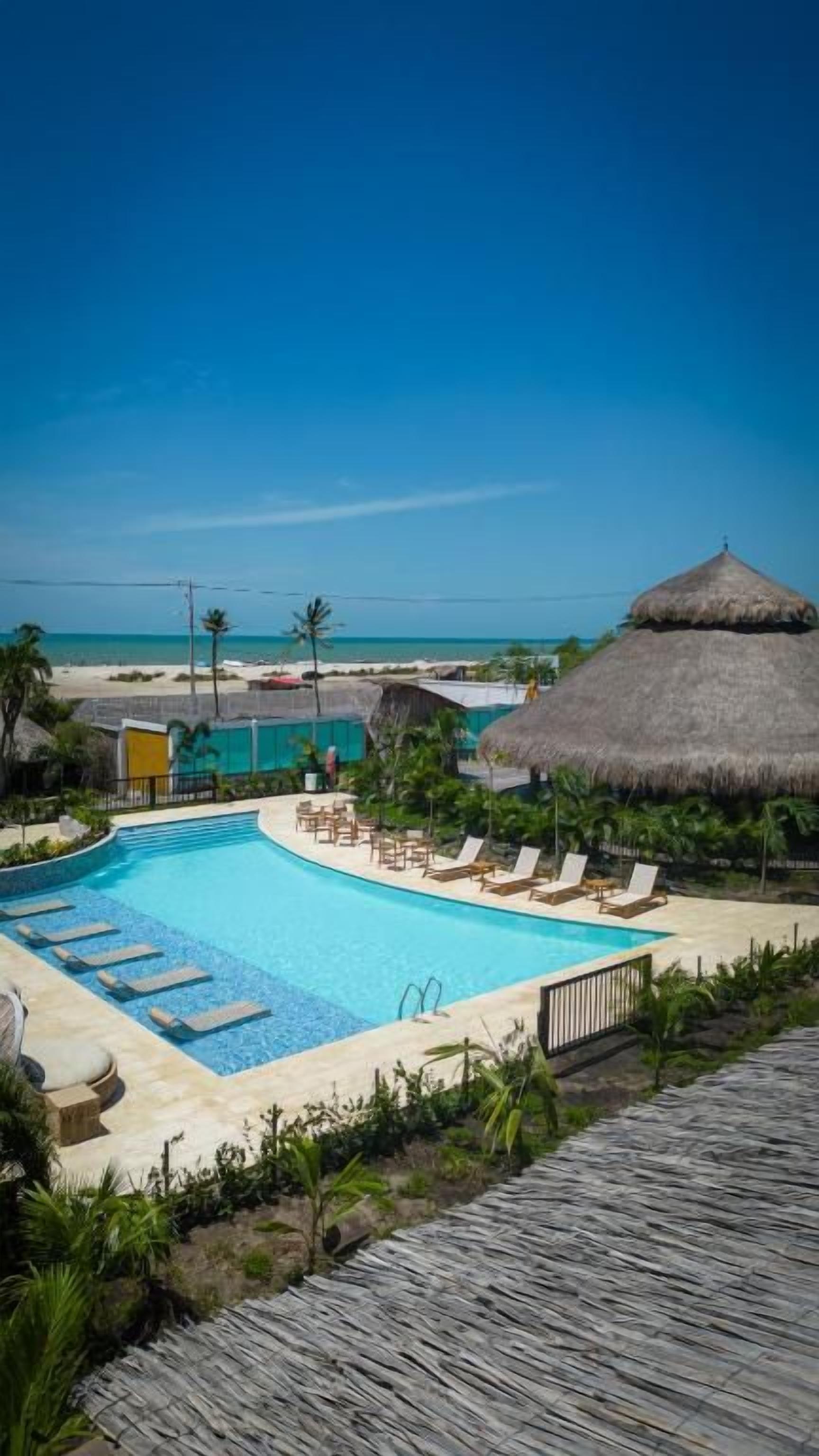 A swimming pool with lounge chairs, a thatched-roof structure, and a view of the beach.