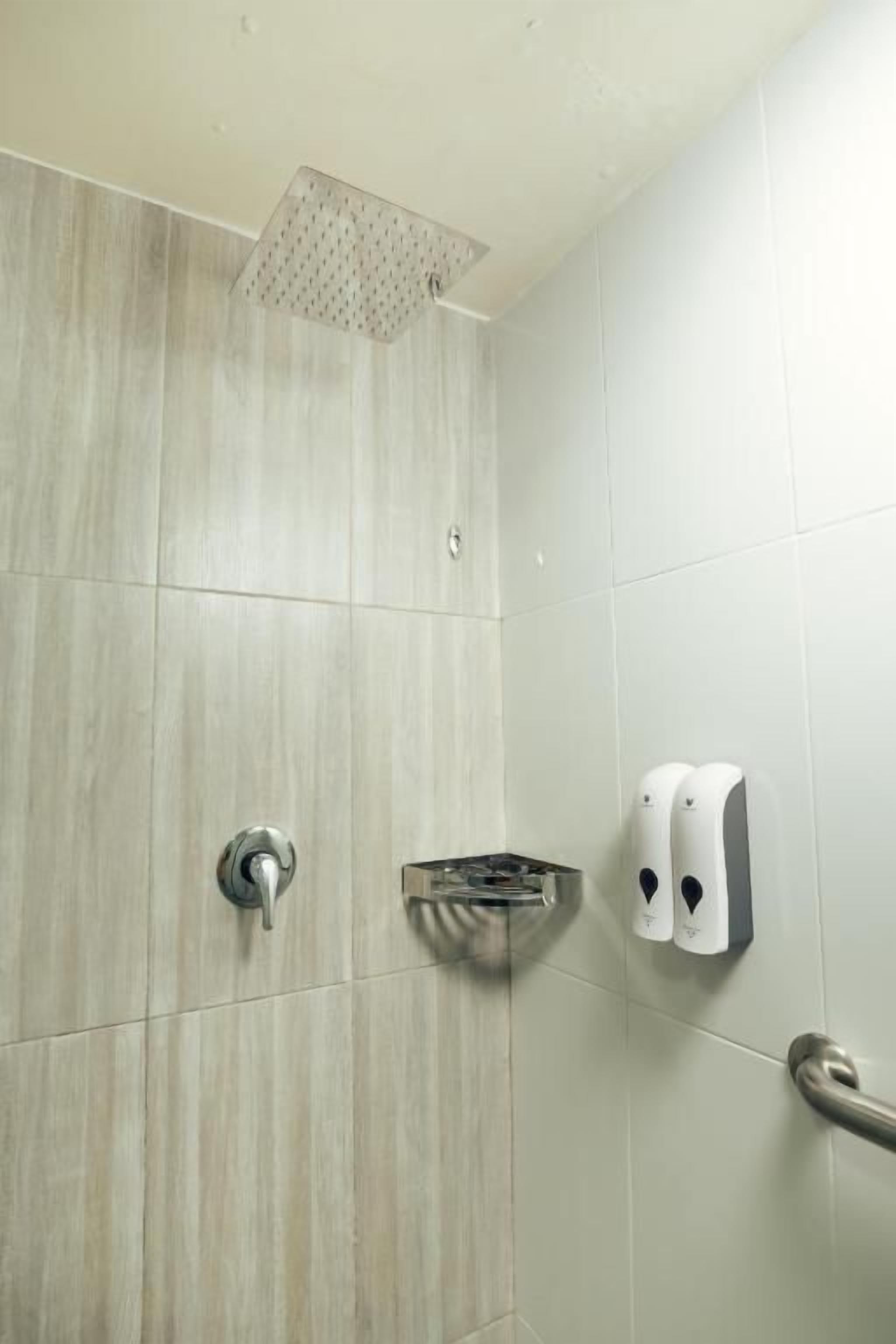 A shower area with a square showerhead, a hand soap dispenser, and a grab bar on the wall.