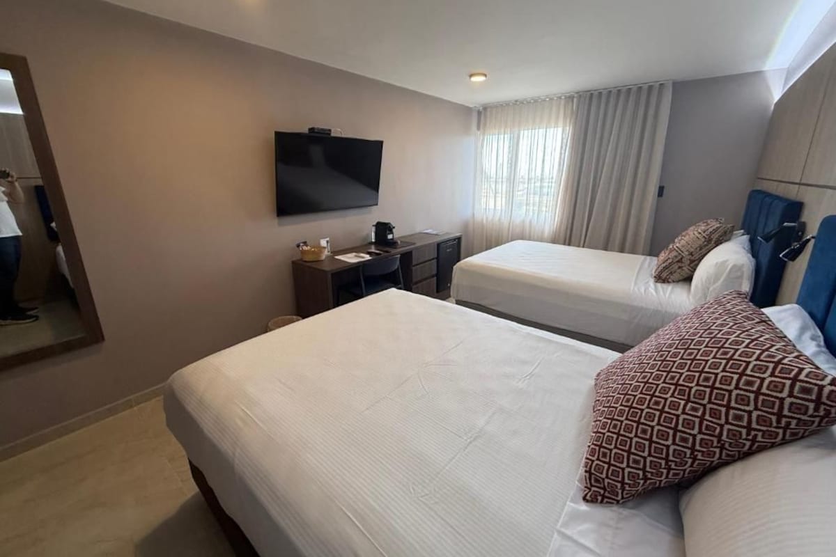 Une chambre d’hôtel avec deux lits, un téléviseur à écran plat, un bureau avec une lampe et une fenêtre avec des rideaux.
