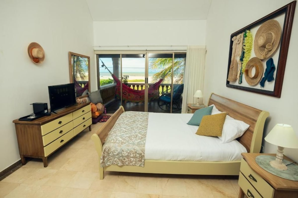 Une chambre avec un lit, une télévision, un bureau et une vue sur la plage, accessible par une porte coulissante.