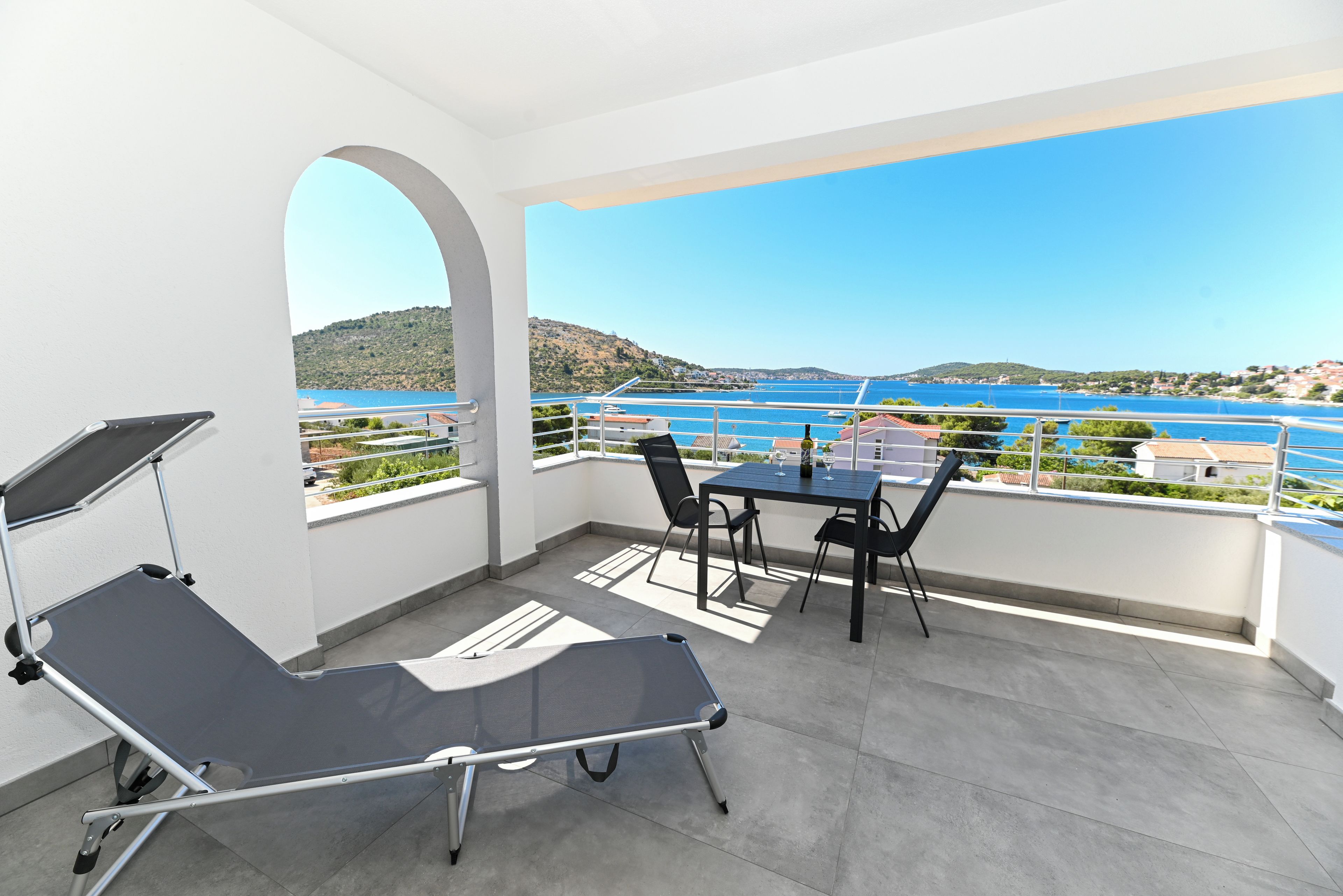 A balcony with a table and chairs, a reclining chair, and a view of a body of water and hills.