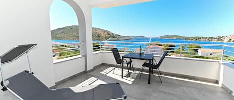 A balcony with a table and chairs, a reclining chair, and a view of a body of water and hills.