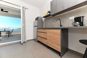 A modern kitchen with a built-in oven, a coffee maker, and a view of the sea through a sliding door. - Lity Studio Gray (Podglavica)