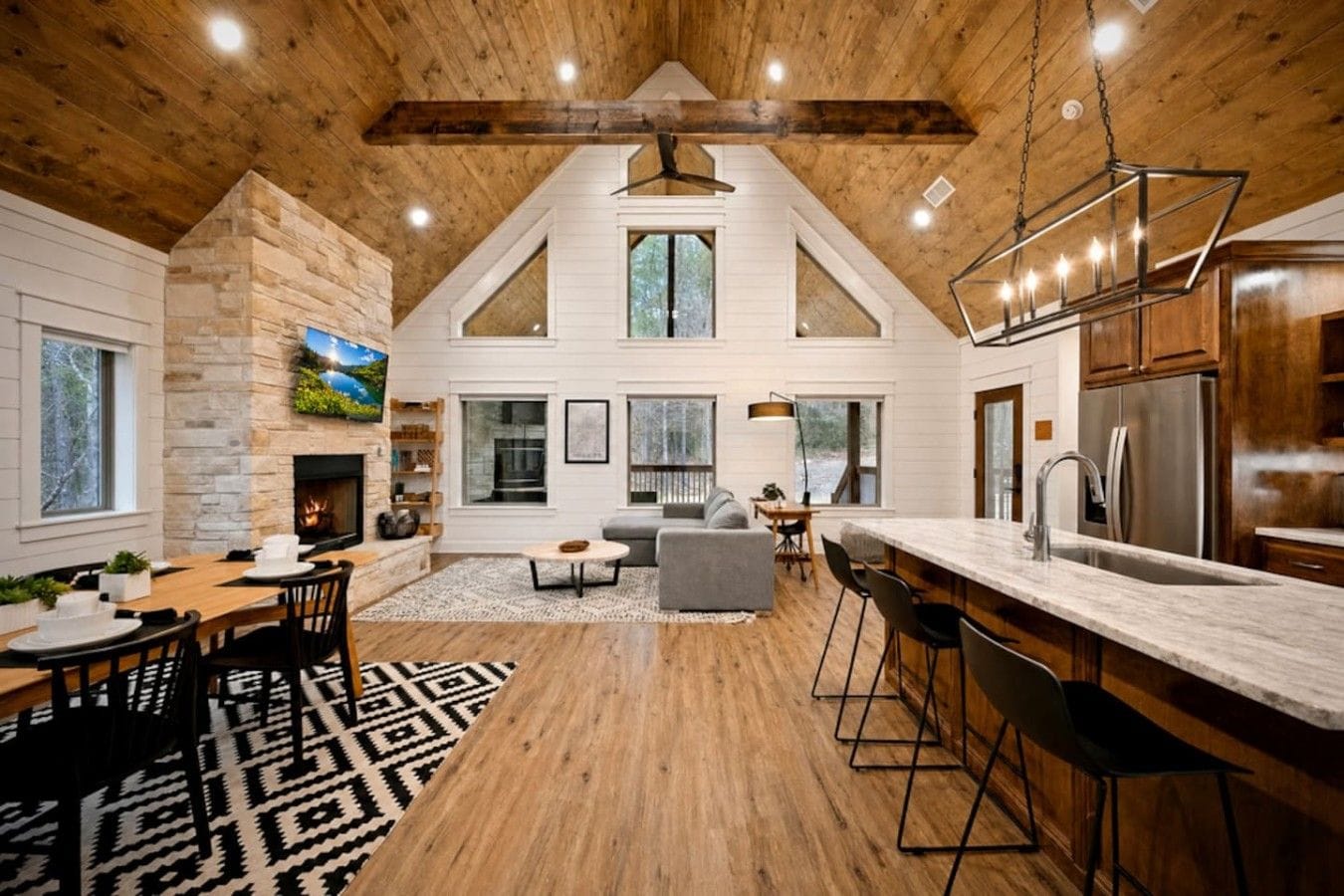 A spacious loft-style interior with a high ceiling, exposed wooden beams, a stone fireplace, a dining area, a living room with a gray sofa, and a kitchen with stainless steel appliances.