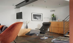 Una sala moderna con un sofá, un sillón reclinable, un televisor de pantalla plana, un cuadro enmarcado en la pared y una escalera.
