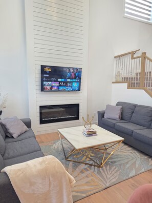 A living room with a flat-screen TV mounted on a wall, a gray sofa, a coffee table with books, and a staircase with wooden railings. - Fort Casamingo 3 bedroom, 3 full bath beautiful home/ Free Wifi (Fort Saskatchewan)