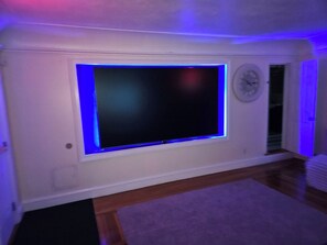 A room with a large flat-screen TV mounted on a white wall, a clock, and a wooden floor. - BIG POOL!!! HOT TUB 5 bed 3 bath 3 min downtown (Providence)
