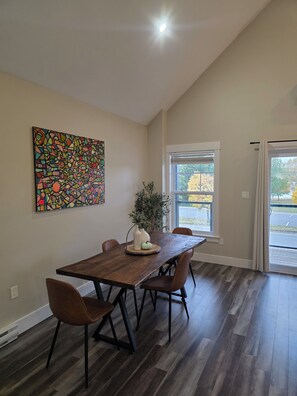 Een eetgedeelte met een houten tafel, vier stoelen, een potplant en een kleurrijk abstract schilderij aan de muur.