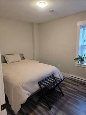 Een slaapkamer met een bed, een bankje, een raam met een plant en een plafondlamp.