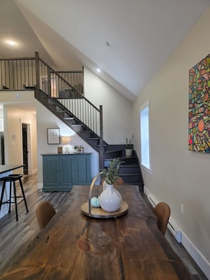 Amplia sala de estar con una mesa de comedor de madera, una escalera y una cocina al fondo.