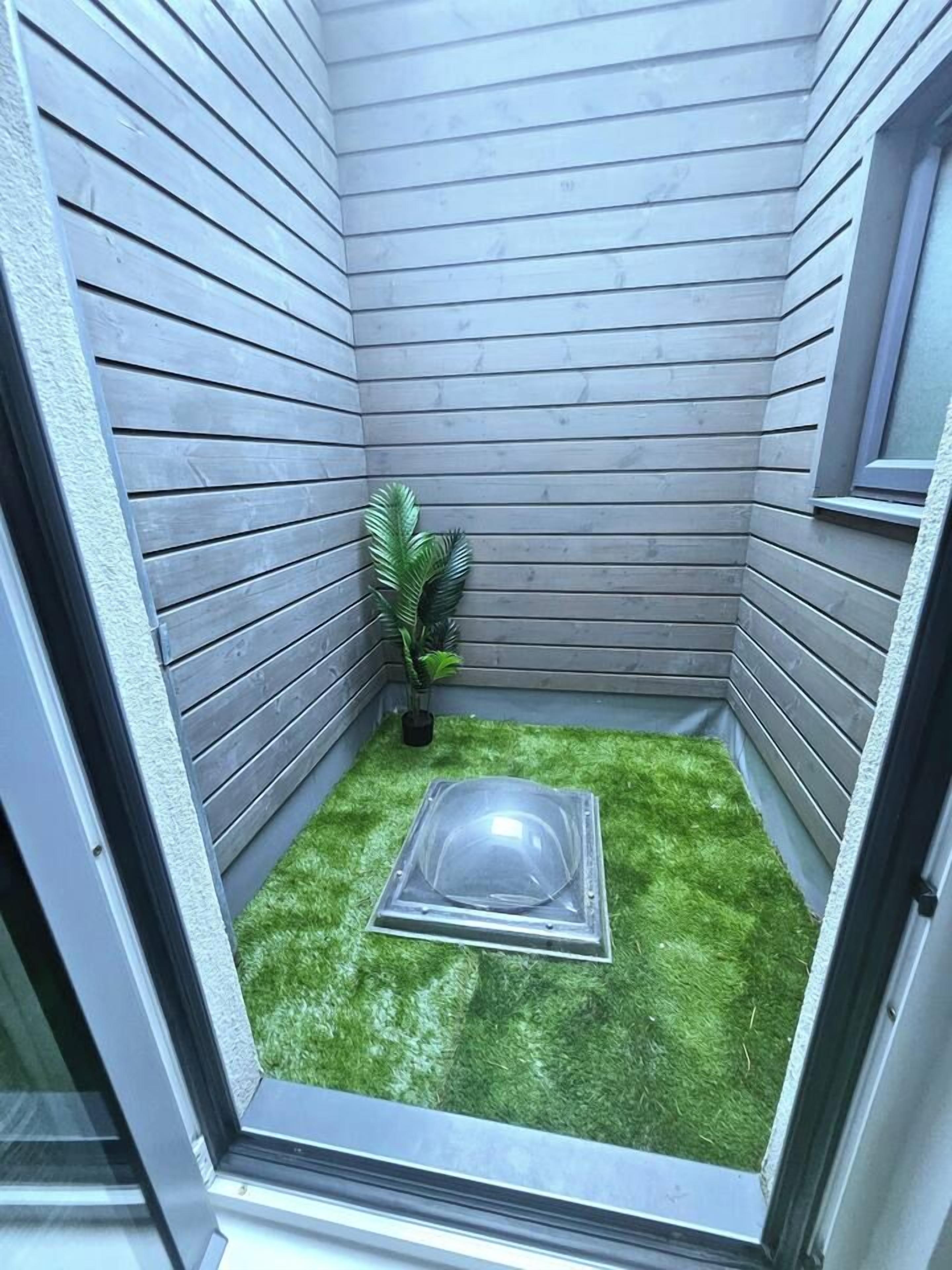 Un petit espace clos doté d’une fenêtre, d’un revêtement de sol en gazon synthétique, d’un objet transparent et d’une plante en pot.
