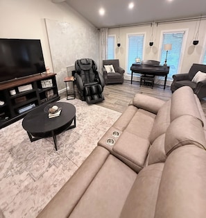 Una sala moderna con un sofá seccional, un sillón de masaje, un televisor de pantalla plana, un piano y una mesa de centro.