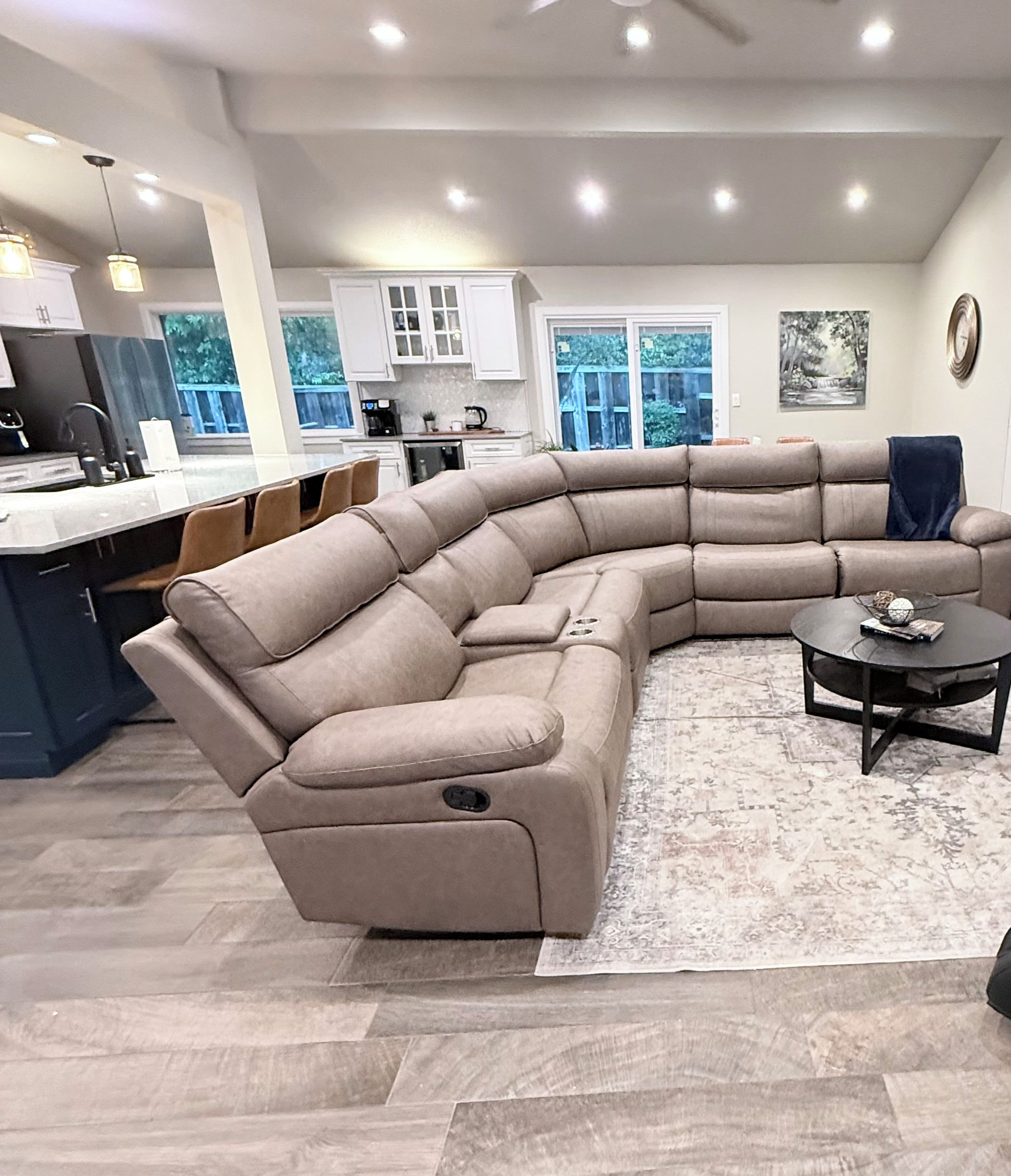 Una sala moderna con un amplio sofá seccional en forma de L, un comedor y una cocina al fondo, con vistas al exterior a través de grandes ventanales.