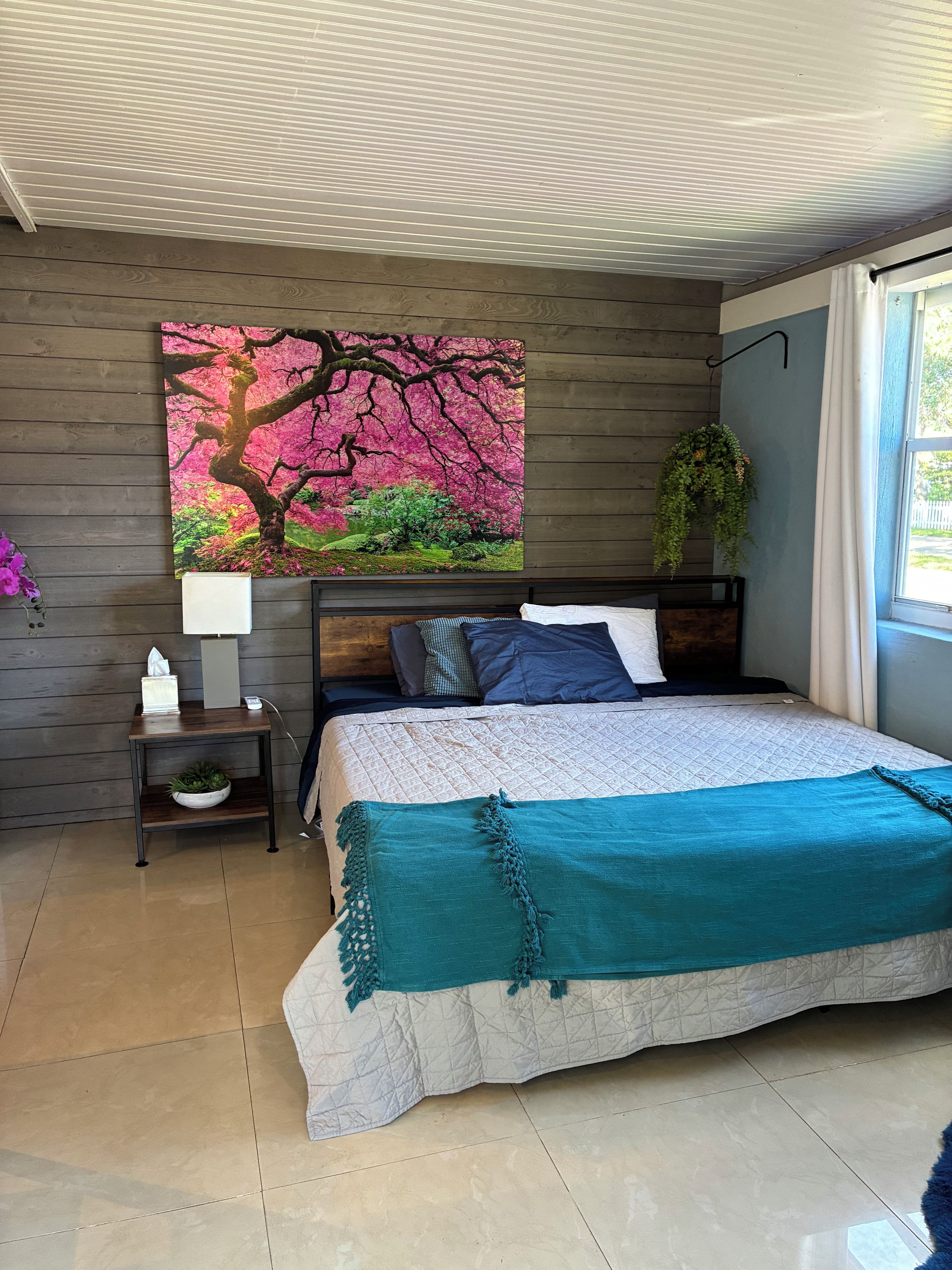 A bedroom with a bed, a nightstand, a wall-mounted painting, a hanging plant, and a window.