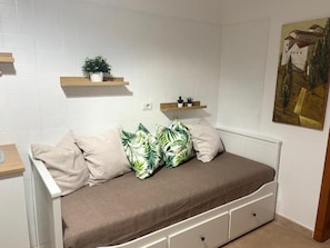 A small, white-framed sofa bed with a brown cover and decorative pillows, flanked by wooden shelves with potted plants, against a white tiled wall. - New Listing (Bari)