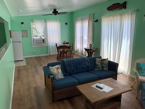 Una sala de estar con sofá, mesa de centro, mesa de comedor y sillas. Hay una zona de cocina con refrigerador y ventilador de techo.