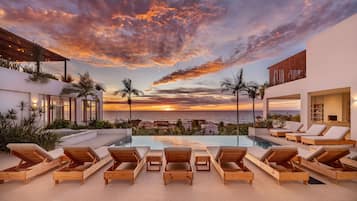 Área da piscina com espreguiçadeiras, vista para o oceano e um pôr do sol.