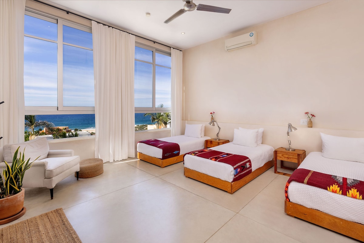 Ein Hotelzimmer mit zwei Betten, einem Sessel, einer Zimmerpflanze und Blick auf den Ozean.