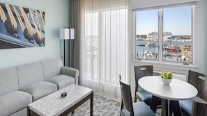 Una habitación de hotel moderna con un sofá gris, una mesa de centro de mármol, una mesa de comedor con sillas, un ventanal con vistas a un puerto deportivo y barcos, y un cuadro en la pared.