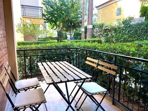 Ein Balkon mit Holztisch und zwei Stühlen, umgeben von einem Metallgeländer und Grünpflanzen.