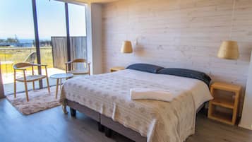 Un dormitorio con una cama grande, piso de madera, una mesita, dos sillas y vistas a través de un ventanal.