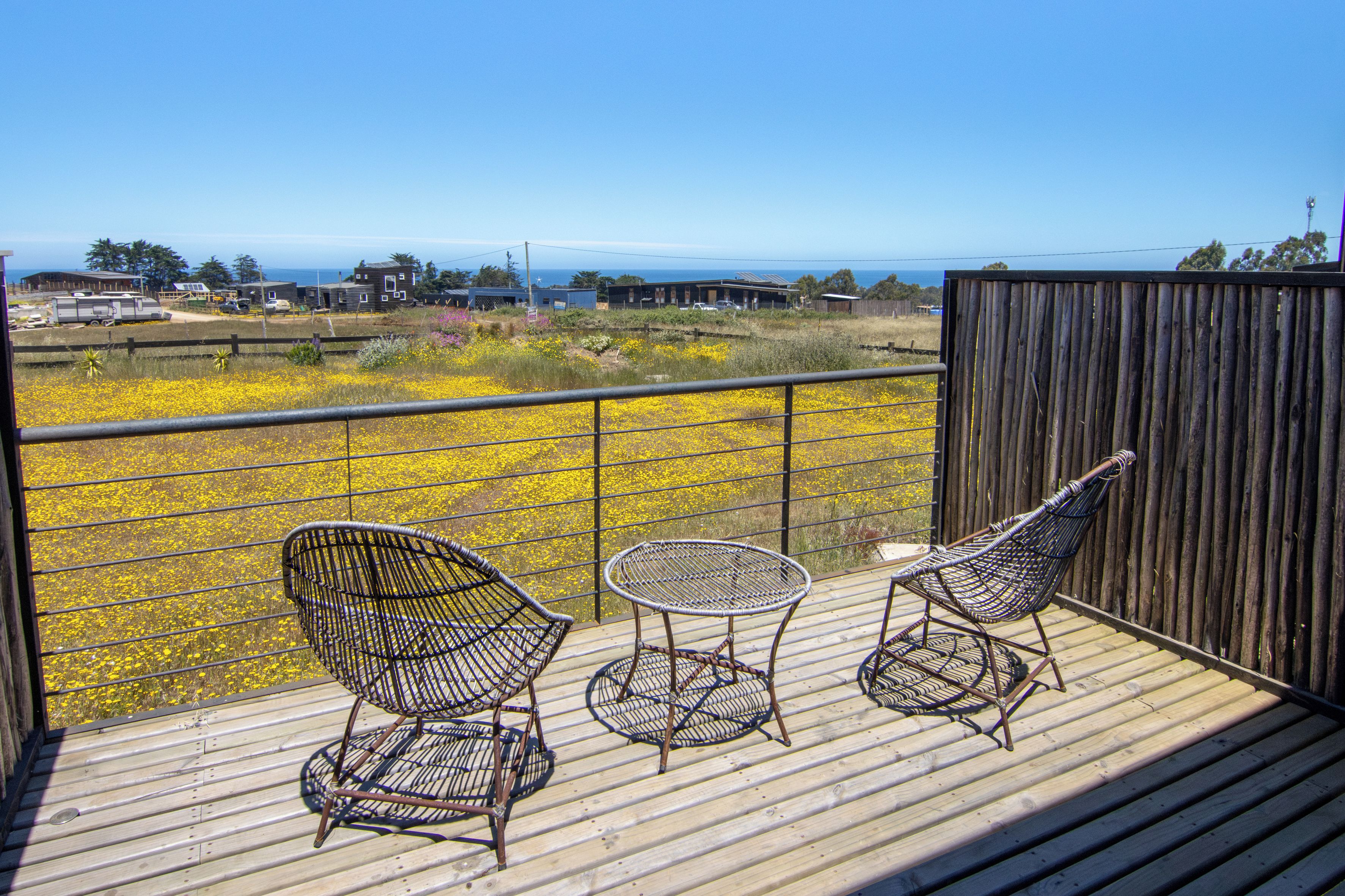 Una terraza de madera con tres sillas de mimbre y una mesita, con vistas a un campo de flores amarillas y una lejana panorámica de casas y el océano.