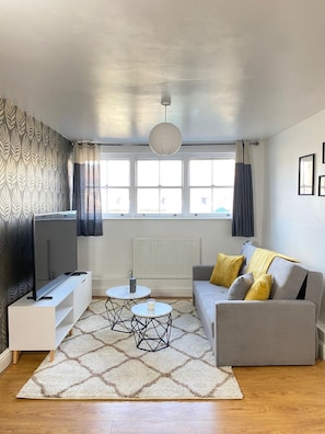 A modern living room with a flat-screen TV, a grey sofa with yellow cushions, a patterned rug, and a window with curtains. - Luxury Brick Lane Flat | Sleep 6 (London)