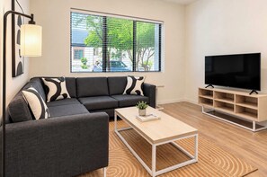 A modern living room with a sectional sofa, a coffee table, a TV stand, and a window with blinds. - Boulevard Stays Modern Tempe 3BR ASU Pool, Gym (Tempe)