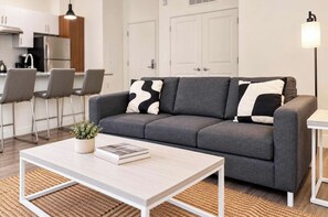 A modern living room with a grey sofa, two black and white pillows, a white coffee table with a book and a plant, and a floor lamp. - Boulevard Stays Modern Tempe 2BR ASU Pool, Gym (Tempe)