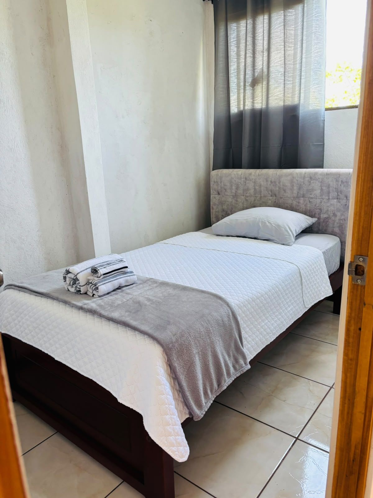 Ein kleines Zimmer mit einem Einzelbett, einem hölzernen Kopfteil, einer grauen Decke und einem gefalteten Handtuch auf dem Bett.