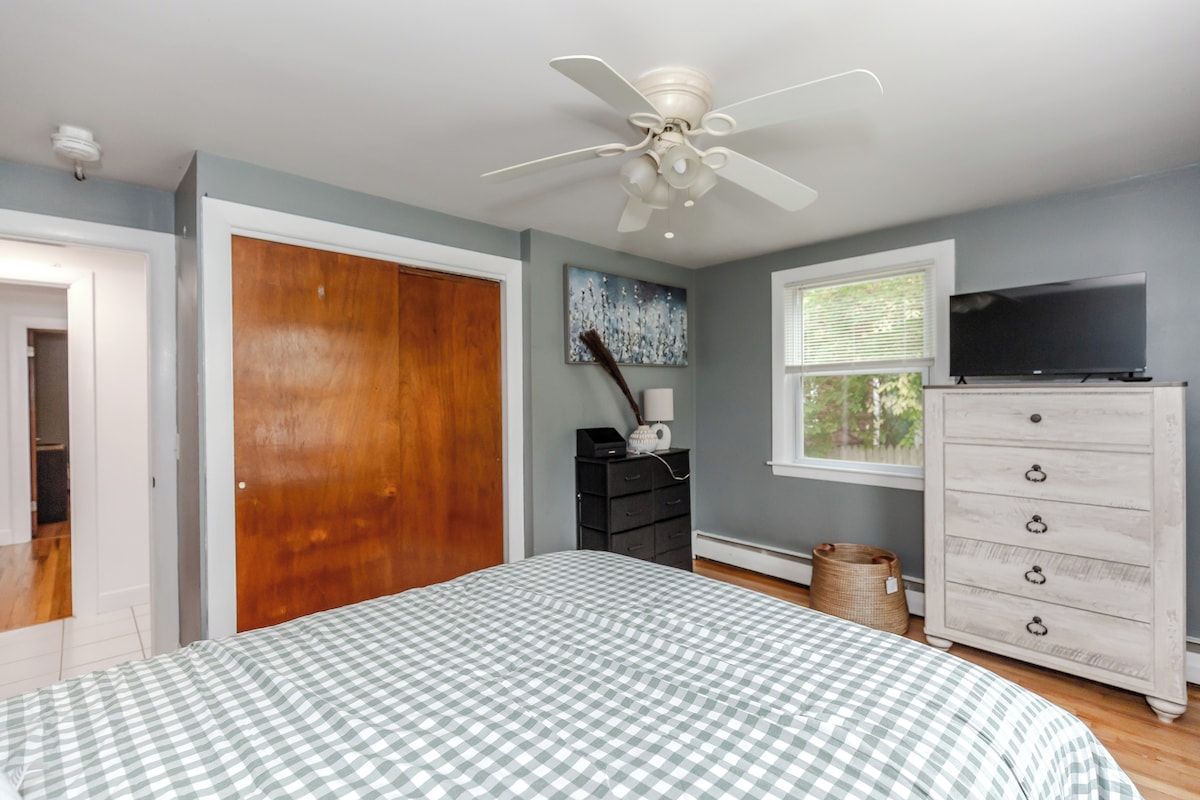 Une chambre à coucher comprenant un lit, un ventilateur de plafond, une commode, un téléviseur et une fenêtre.
