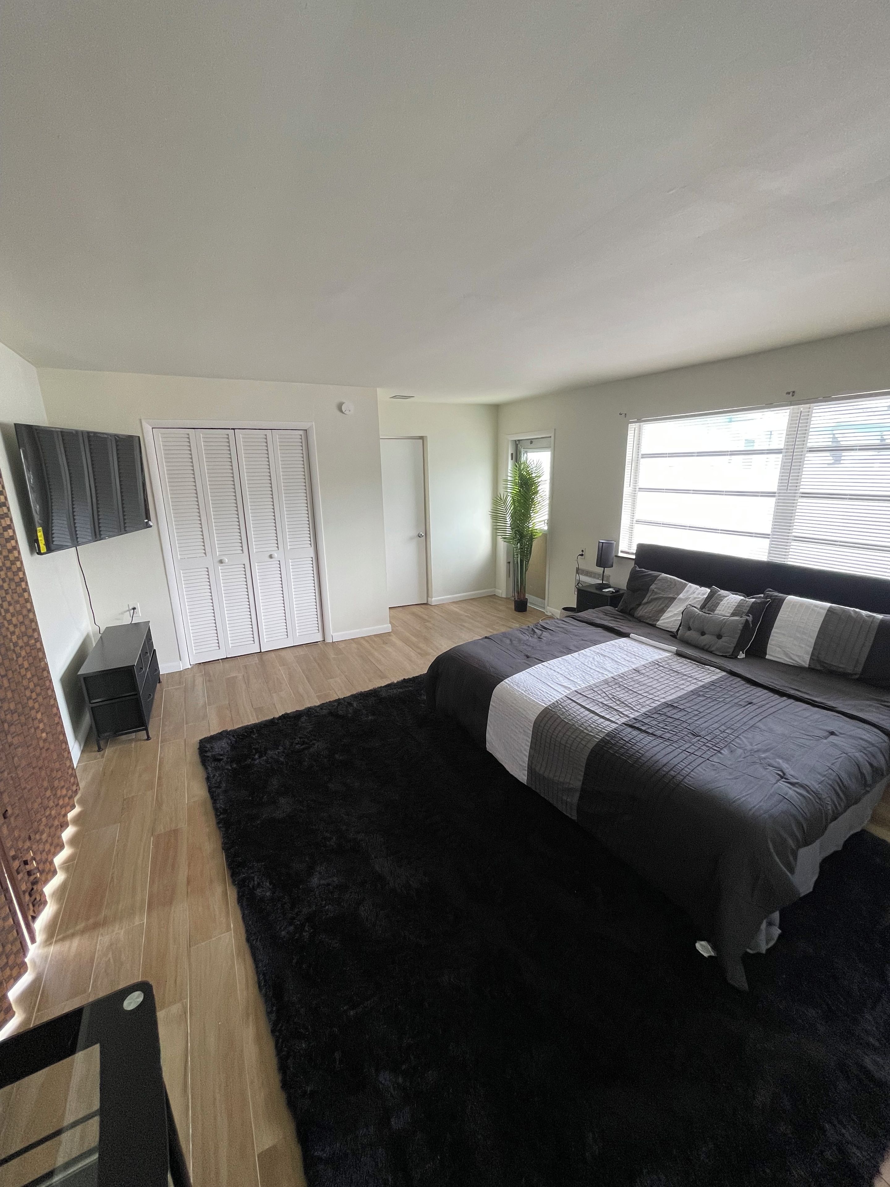 Ein Schlafzimmer mit einem großen Bett, einem schwarzen Teppich, einem Schminktisch, einem Fernseher und einem Fenster mit Jalousien.