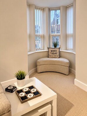 A corner room with a curved bench, a small table with a plant and a game, and a window with a view of buildings. - Stylish Central Lytham 2 Bed - Walk Everywhere (Lytham Saint Annes)