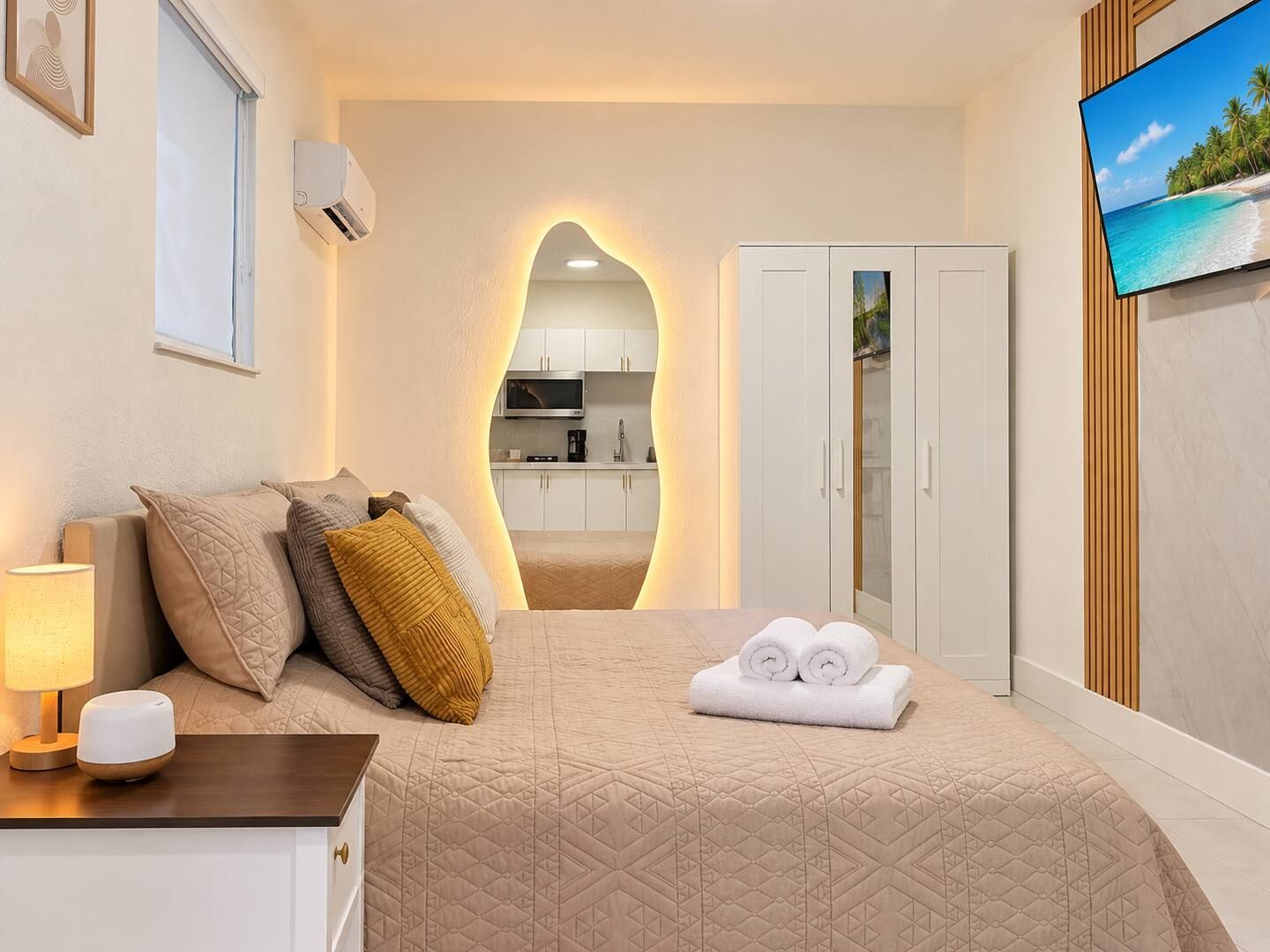 Habitación de hotel con cama, televisor, escritorio con lámpara, armario y vista a la cocina.