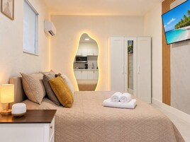 Habitación de hotel con cama, televisor, escritorio con lámpara, armario y vista a la cocina.