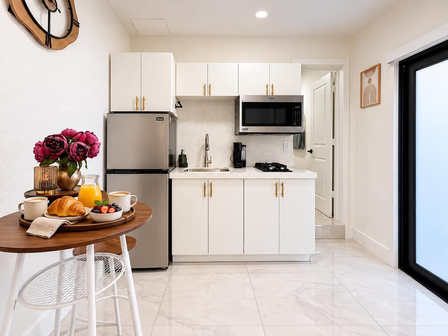 Una cocina moderna con armarios blancos, un refrigerador de acero inoxidable, un microondas y una cafetera. Hay un desayuno listo con croissants, fruta y una taza de café sobre una mesa redonda.
